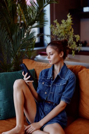 Téléchargez les photos : Jeune femme assise sur un canapé, absorbée dans son téléphone portable, absorbée dans le monde numérique - en image libre de droit
