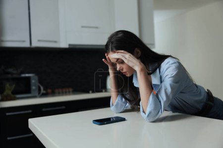 Foto de Mujer estresada sentada en el mostrador de la cocina con teléfono delante de ella, sintiéndose abrumada y exasperada mientras sostiene su cabeza en las manos - Imagen libre de derechos