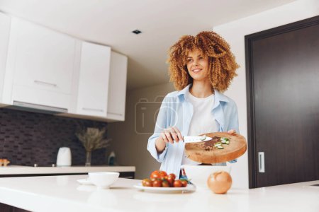 Photo for A woman with curly hair preparing fresh vegetables on a wooden cutting board in the kitchen - Royalty Free Image