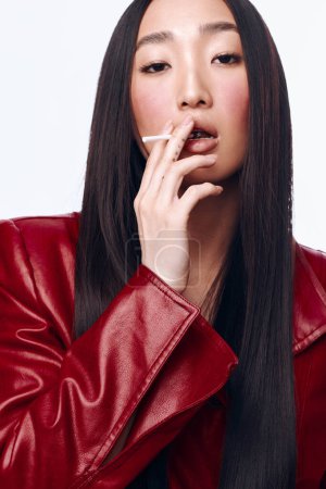 Foto de Mujer asiática con estilo en chaqueta de cuero rojo fumar cigarrillo sobre fondo blanco con expresión de confianza - Imagen libre de derechos
