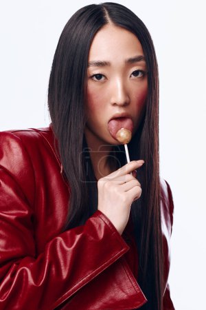Foto de Elegante mujer asiática en chaqueta de cuero rojo con piruleta en la boca contra fondo gris - Imagen libre de derechos