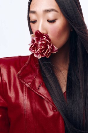 Foto de Hermosa mujer en chaqueta de cuero rojo con los ojos cerrados sosteniendo flor en la boca retrato al aire libre - Imagen libre de derechos