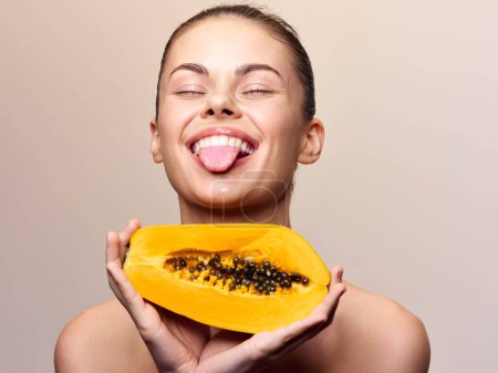 Téléchargez les photos : Femme ludique tenant tranche de papaye avec la langue dehors, appréciant les fruits tropicaux par une journée ensoleillée - en image libre de droit