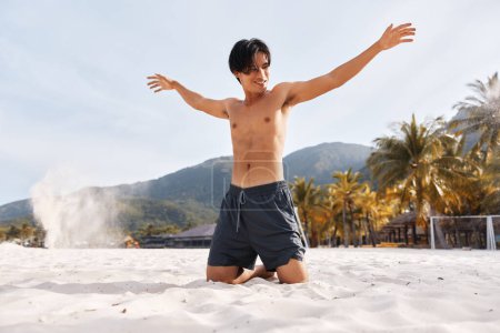 Hombre asiático sonriente disfrutando de vacaciones en la playa, abdomen expuesto, torso muscular, fondo de palma tropical