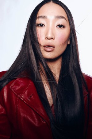 Téléchargez les photos : Femme asiatique élégante avec cheveux noirs en veste en cuir rouge et rouge à lèvres posant pour appareil photo - en image libre de droit