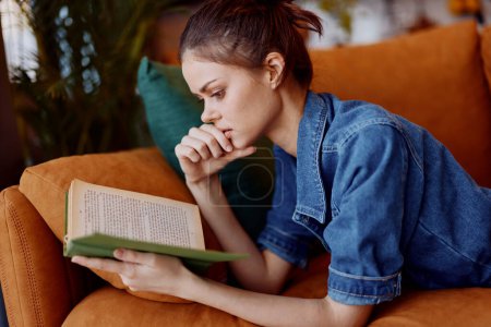 Téléchargez les photos : Femme se détendre et lire un livre sur un canapé confortable dans un salon confortable avec une plante verte en arrière-plan - en image libre de droit