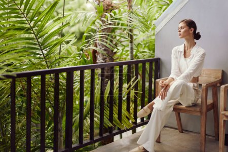Smiling Woman Enjoying Tropical Vacation on Balcony with a Stunning View A happy woman stands on a sunny balcony, surrounded by lush greenery and a breathtaking tropical landscape She wears trendy