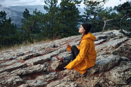 Photo for Woman in a yellow raincoat enjoying the serene beauty of nature, sitting on a rock in front of a majestic mountain landscape - Royalty Free Image