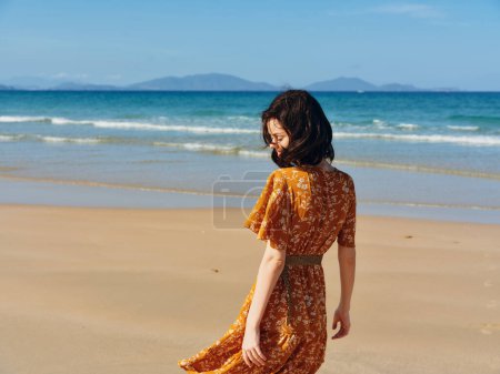 Photo for Woman in an orange dress strolling along the shoreline of a calm beach with a clear blue ocean in the background - Royalty Free Image