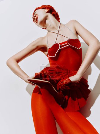 Foto de Mujer con estilo en vestido rojo y tacones altos posando con confianza en la superficie blanca con las manos en las caderas - Imagen libre de derechos