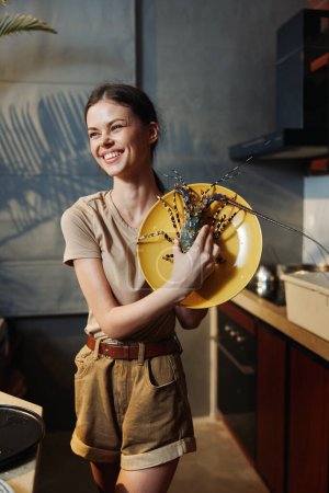 Téléchargez les photos : Femme heureuse tenant une assiette avec un délicieux homard cuit dans la cuisine confortable à la maison - en image libre de droit