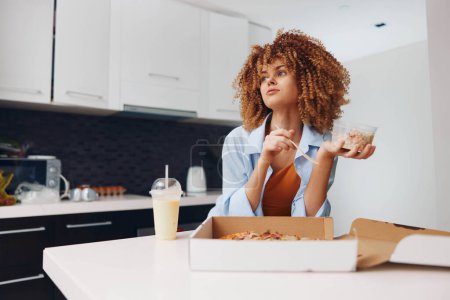 Téléchargez les photos : Femme aux cheveux bouclés appréciant la pizza tout en restant debout dans la cuisine tenant la boîte à pizza - en image libre de droit