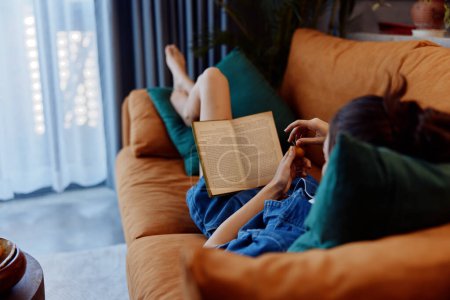 Téléchargez les photos : Femme relaxante sur un canapé près de la fenêtre, absorbée par un livre dans son confortable salon à la maison - en image libre de droit