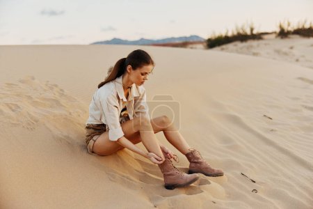 Lonely woman sitting in sandy desert with boots on her feet under the scorching sun, looking for direction and solace