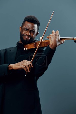 Photo for Elegant African American man in black suit playing violin on gray background in dramatic lighting - Royalty Free Image