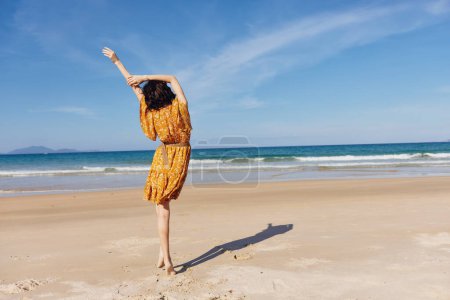Femme en robe jaune debout avec les bras tendus sur la plage de sable sous les rayons du soleil