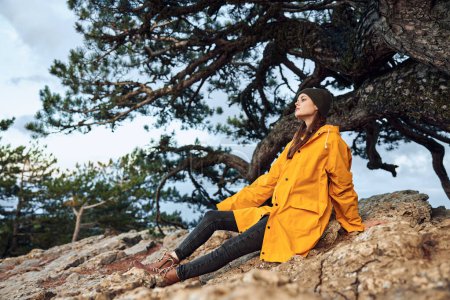 Foto de Mujer joven y serena en impermeable amarillo disfrutando de la naturaleza en una roca cerca de un pino en una escena de belleza de aventura de viaje salvaje - Imagen libre de derechos