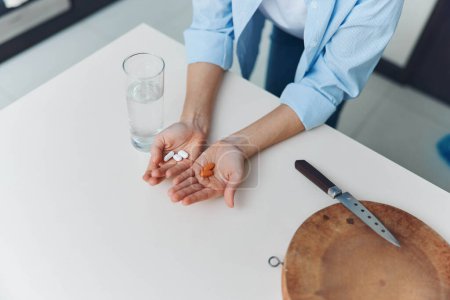 Person hält Tabletten neben Messer und Glas Wasser auf Tisch