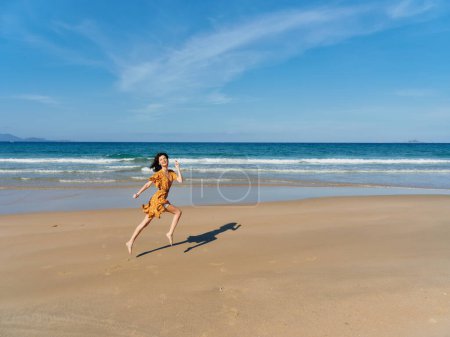 Téléchargez les photos : Femme heureuse courir en robe jaune sur la plage de sable avec fond bleu ciel clair - en image libre de droit