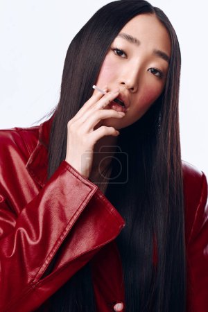 Téléchargez les photos : Femme asiatique élégante fumant une cigarette en cuir rouge veste avec de longs cheveux noirs portrait - en image libre de droit