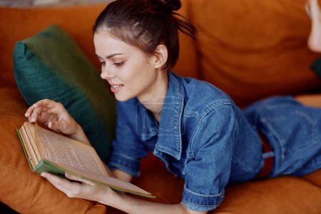Téléchargez les photos : Femme relaxante sur le canapé à la maison, livre de lecture en chemise denim, scène intérieure confortable et confortable - en image libre de droit