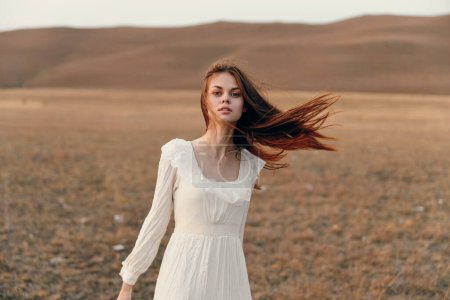 Foto de Mujer en vestido blanco de pie en el campo ventoso rodeado de naturaleza y libertad belleza y serenidad en la naturaleza - Imagen libre de derechos