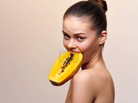 Téléchargez les photos : Attractive female model holding a fresh papaya fruit in her mouth, on a neutral beige background - en image libre de droit