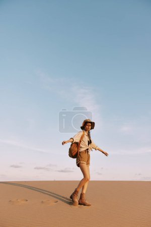Woman in hat and shorts exploring the vast desert landscape, walking across a sandy dune under the scorching sun
