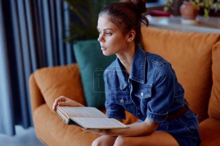 Photo for Woman in denim outfit reading book on couch in cozy living room setting - Royalty Free Image