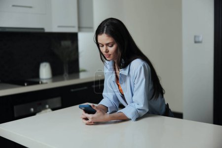Téléchargez les photos : Femme assise sur le comptoir de la cuisine la nuit, absorbée dans l'écran du téléphone, créant une atmosphère tranquille et confortable - en image libre de droit
