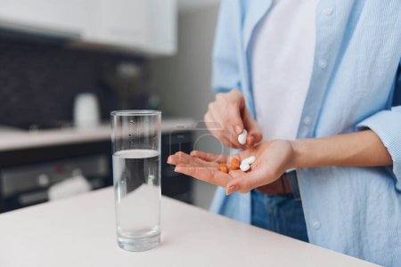 Téléchargez les photos : Femme tenant des pilules et un verre d'eau sur le comptoir dans un intérieur de cuisine moderne avec lumière naturelle - en image libre de droit