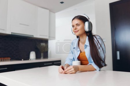Foto de Young woman enjoying a cup of coffee while listening to music with headphones at the kitchen table - Imagen libre de derechos
