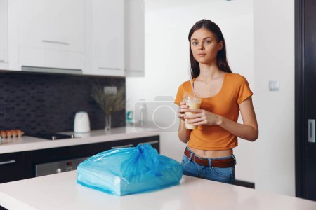 Frau steht mit Tasse Kaffee und Müllsack in Küche, Hausarbeit-Konzept