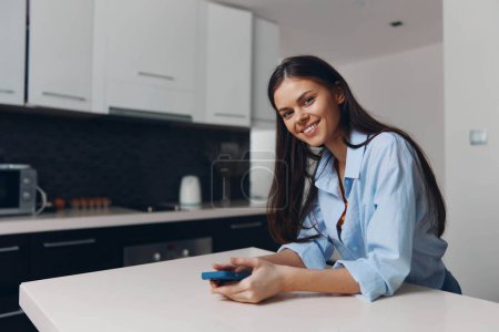 Photo for Smiling woman sitting at kitchen table holding cell phone in hand, looking happy and content - Royalty Free Image