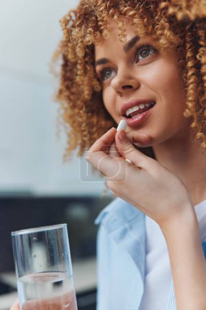 Photo for Curly haired woman taking a pill with a glass of water in hand, health and wellness concept - Royalty Free Image