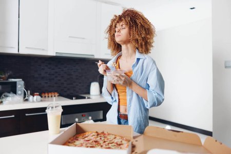 Téléchargez les photos : Femme aux cheveux bouclés dégustant une tranche de pizza tout en se tenant devant une boîte de pizza - en image libre de droit
