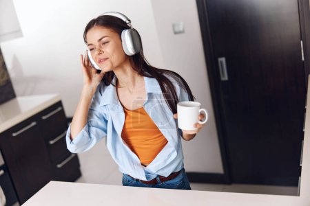 Téléchargez les photos : Jeune femme dans les écouteurs profiter de la musique et du café dans la cuisine tout en vérifiant son téléphone - en image libre de droit