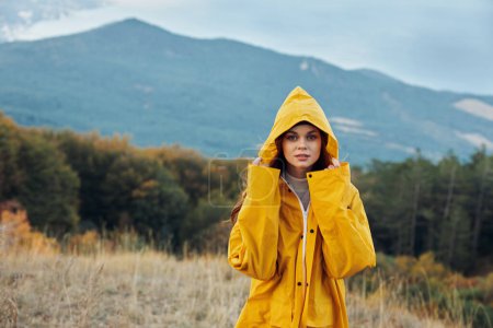 Photo for Woman in yellow raincoat standing in field with mountain backdrop Traveler admiring nature beauty under cloudy sky - Royalty Free Image