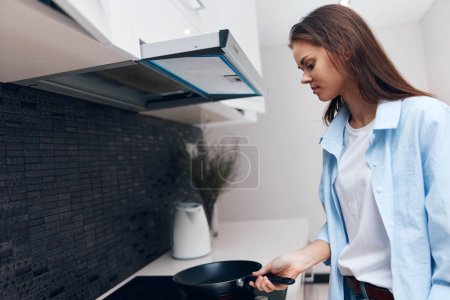 Woman in Blue Shirt Cooking with Frying Pan at Stove in Domestic Kitchen Interior Concept