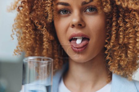 Téléchargez les photos : Femme aux cheveux bouclés tenant un verre d'eau, prenant une pilule dans la bouche, concept de soins de santé - en image libre de droit