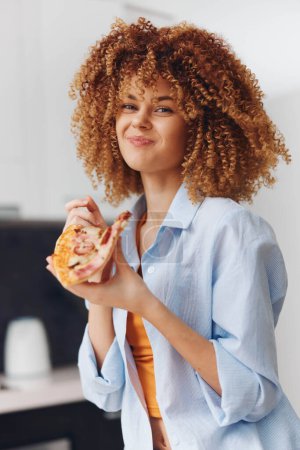 Photo for Young black woman with curly hair enjoying a slice of pizza in her home kitchen, African American female eating delicious food - Royalty Free Image