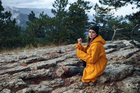 A woman enjoying the serene beauty of nature, sitting on a rock in a yellow coat, surrounded by trees and mountains