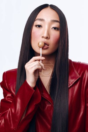 Portrait de jeune femme asiatique élégante avec veste en cuir rouge tenant sucette dans la bouche