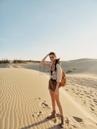 Woman standing in desert with sand dunes in background, carrying backpack on adventurous journey in arid landscape