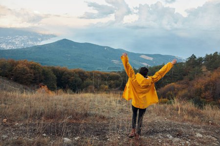 Foto de Una persona en un impermeable amarillo de pie en una colina con los brazos extendidos disfrutando de la belleza de la naturaleza y la libertad - Imagen libre de derechos