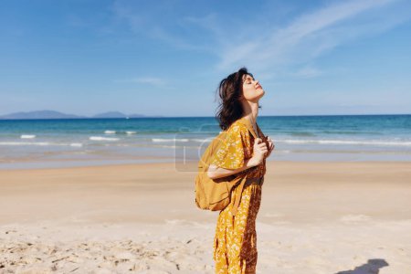 Foto de Mujer excitada en un vibrante vestido amarillo disfrutando de la playa con los brazos levantados en el aire, sintiéndose libre y feliz - Imagen libre de derechos