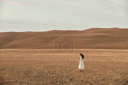 Frau im weißen Kleid steht auf offenem Feld, umgeben von Hügeln, Schönheit der Natur und Gelassenheit Konzept