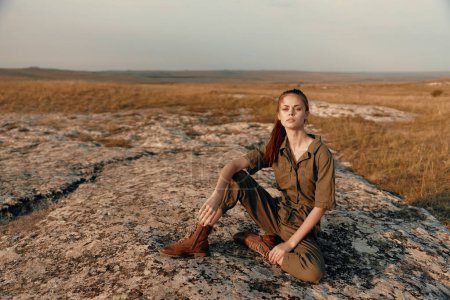 Téléchargez les photos : Jeune femme assise sur le rocher dans le champ avec les mains sur les hanches dans la nature tranquille scène de beauté - en image libre de droit