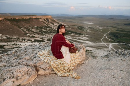 Téléchargez les photos : Femme relaxante sur roche avec couverture à carreaux en face de majestueuse vue sur la montagne pour le voyage et l'aventure concept - en image libre de droit