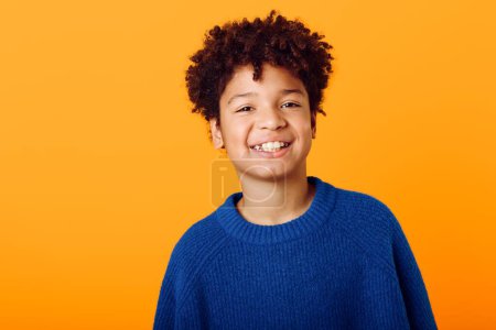 Téléchargez les photos : Lumineux et joyeux un jeune garçon afro-américain joyeux dans un pull bleu sur un fond orange vibrant - en image libre de droit